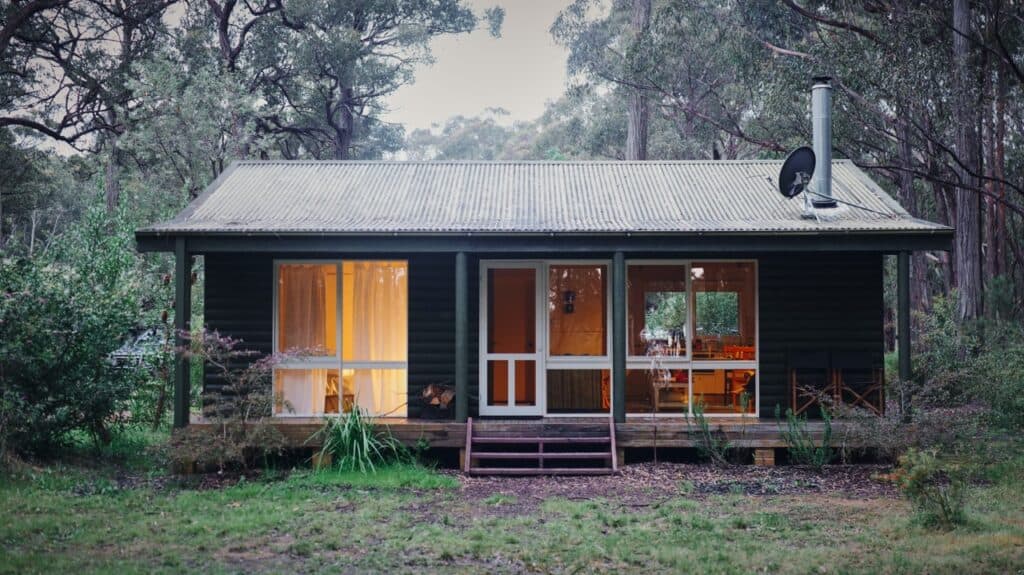 A cozy dark green cabin with large windows and a porch sits surrounded by tall trees and greenery in a Blue Mountains forest setting. Warm light glows from inside, creating an inviting atmosphere perfect for trip planning or following a travel guide. | Megalong Restaurant