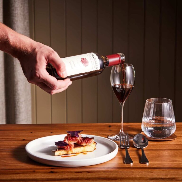 A person pours red wine into a glass beside a plate with a slice of pie, a second empty glass, and cutlery, set on a wooden table with a striped curtain background at one of | Megalong Restaurant