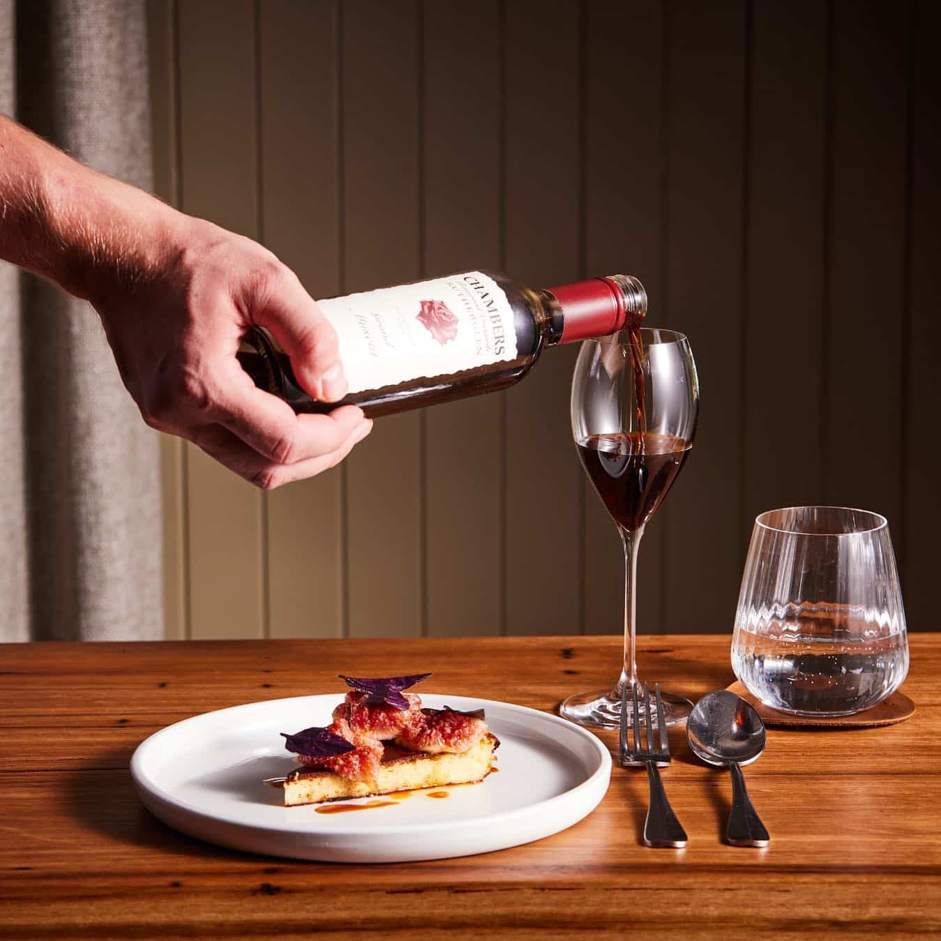 A person pours red wine into a glass beside a plate with a slice of pie, a second empty glass, and cutlery, set on a wooden table with a striped curtain background at one of | Megalong Restaurant
