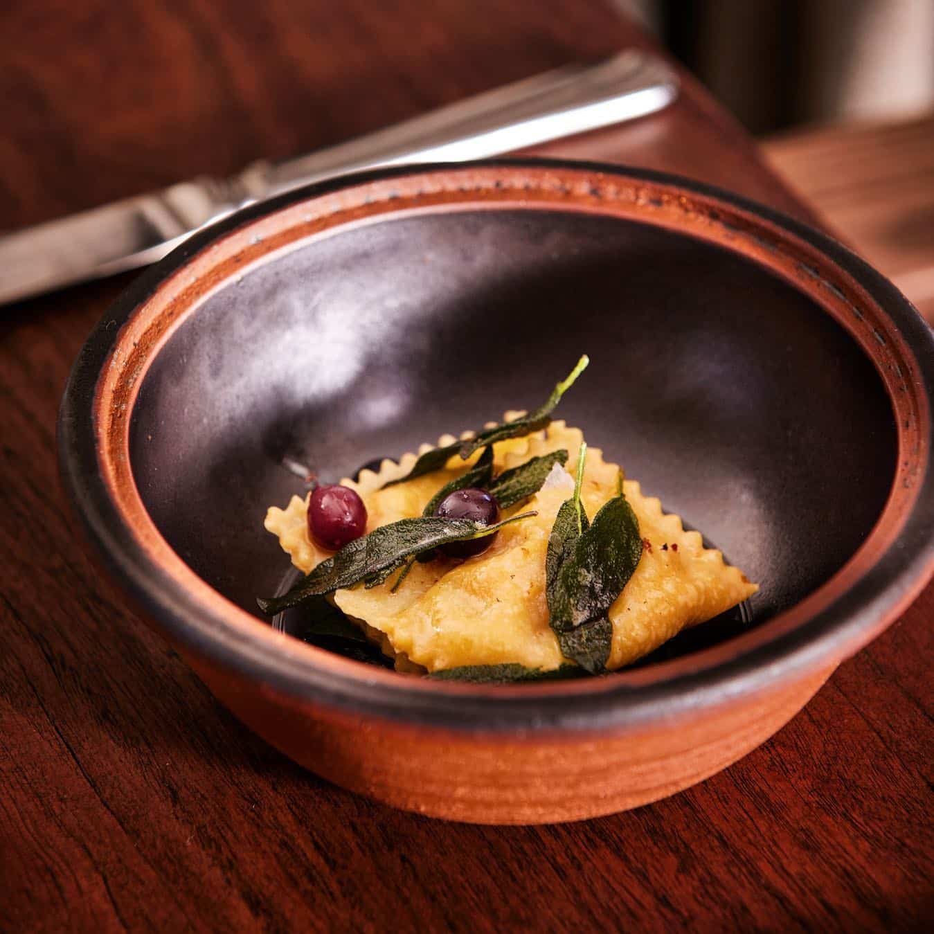 A single ravioli garnished with sage leaves and pomegranate seeds served in a rustic clay bowl, accompanied by a fork on a wooden table at one of the best Blue Mountains restaurants. | Megalong Restaurant