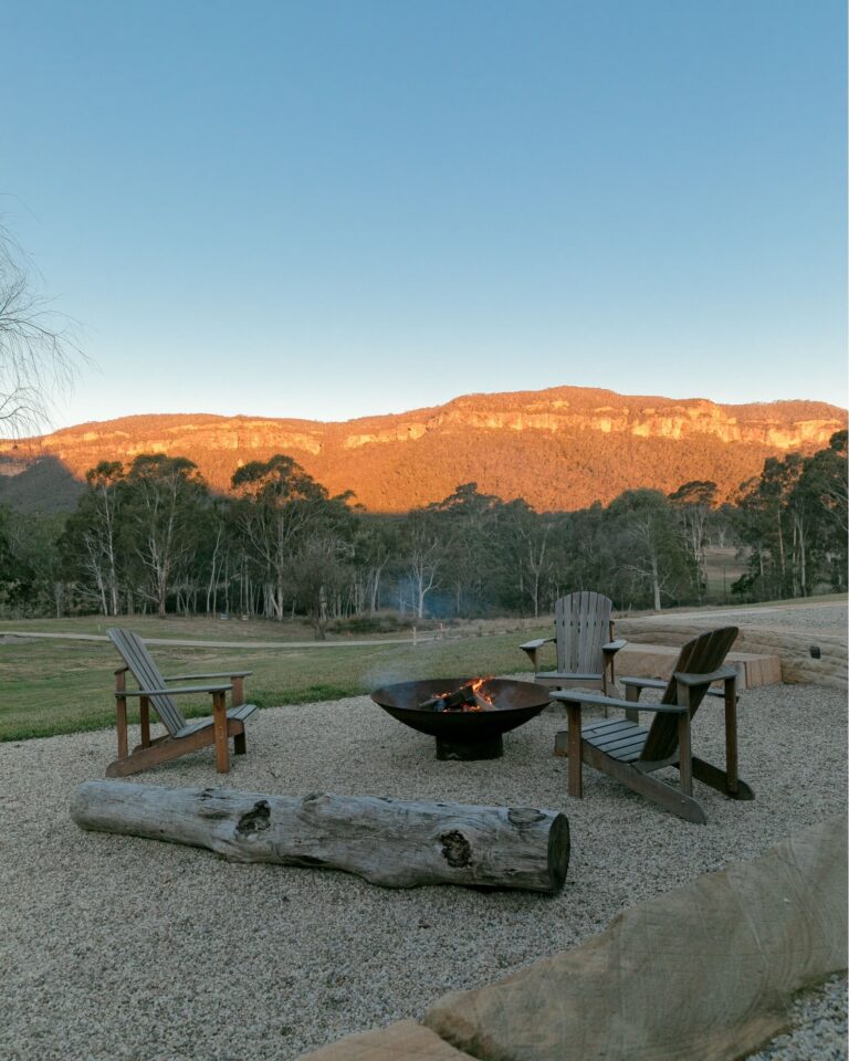 Two wooden chairs facing a burning fire pit in a gravel area with a large log in front, overlooking the scenic Blue Mountains lit by the sunset. | Megalong Restaurant