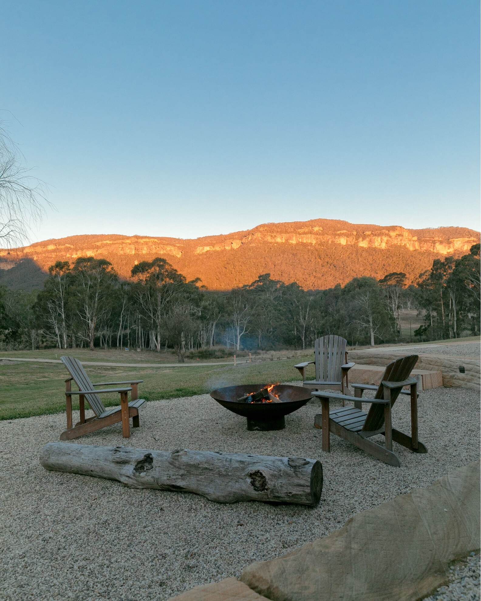 Two wooden chairs facing a burning fire pit in a gravel area with a large log in front, overlooking the scenic Blue Mountains lit by the sunset. | Megalong Restaurant