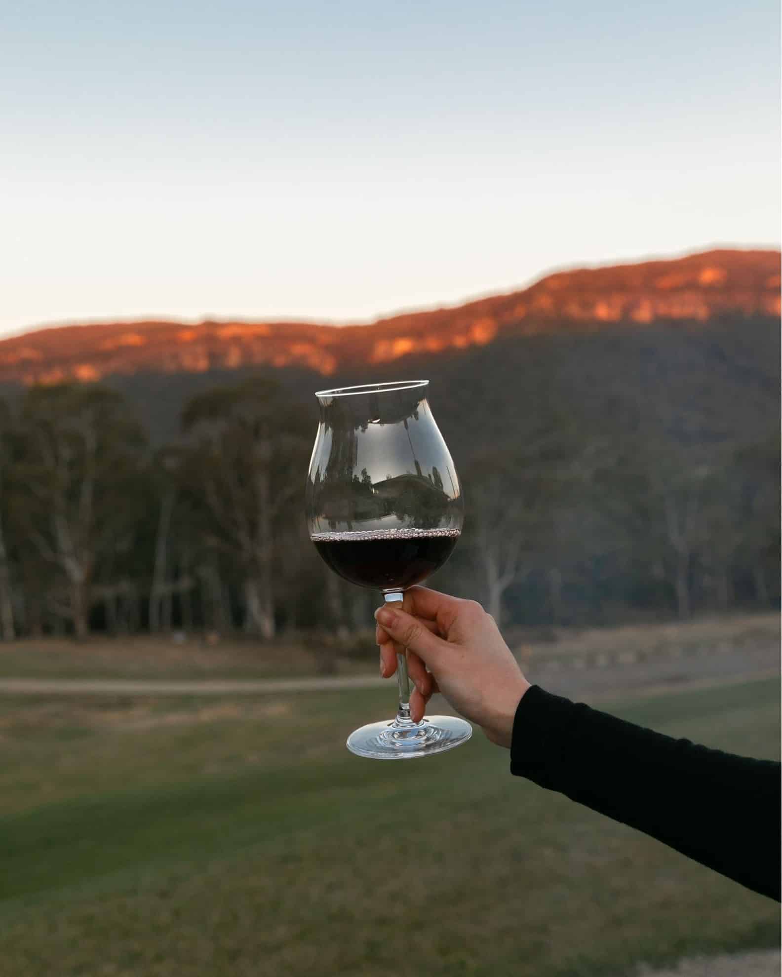 A hand holding a glass of red wine with a scenic background featuring gentle hills and a soft evening sky near the best lunch spots in the Blue Mountains. | Megalong Restaurant