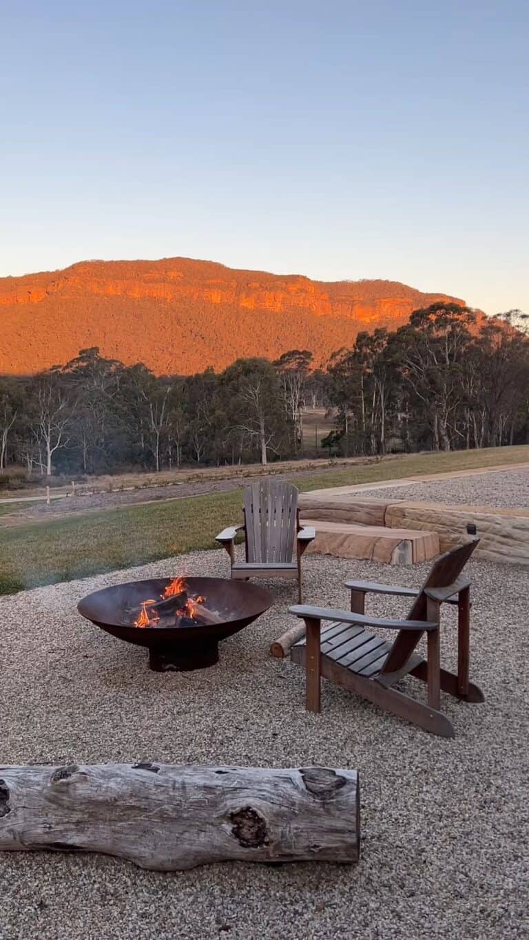 A serene outdoor setting with a fire pit aflame, wooden Adirondack chairs facing the Blue Mountains bathed in golden sunset light, and a tranquil landscape in the background. | Megalong Restaurant
