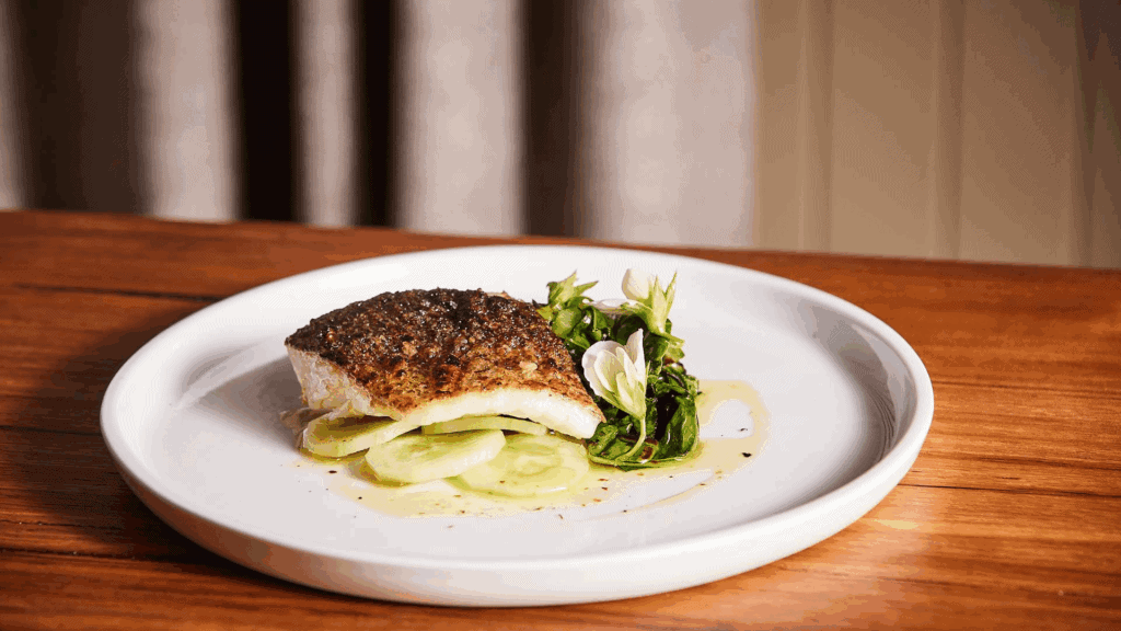 A seared fish fillet with crispy skin served atop thinly sliced cucumbers and a garnish of leafy greens on a white plate, presented on a wooden table with a beige curtain background at | Megalong Restaurant