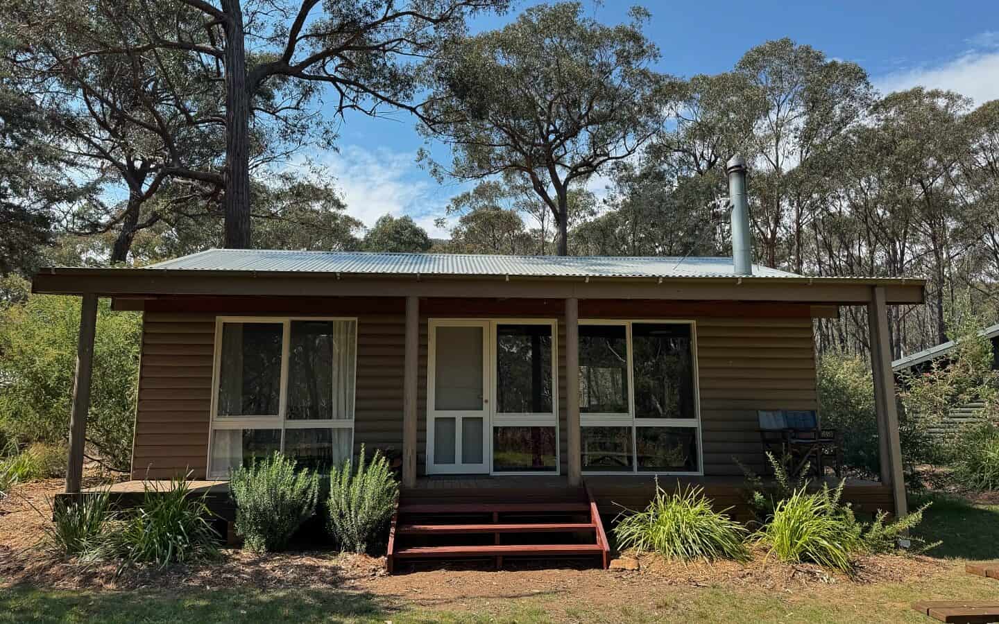 A small brown cabin with a tin roof and a chimney set in a lush, wooded area. Large windows and a white front door are visible, with steps leading up to a small porch. Greenery and tall trees surround the structure under a partly cloudy sky. | Werriberri