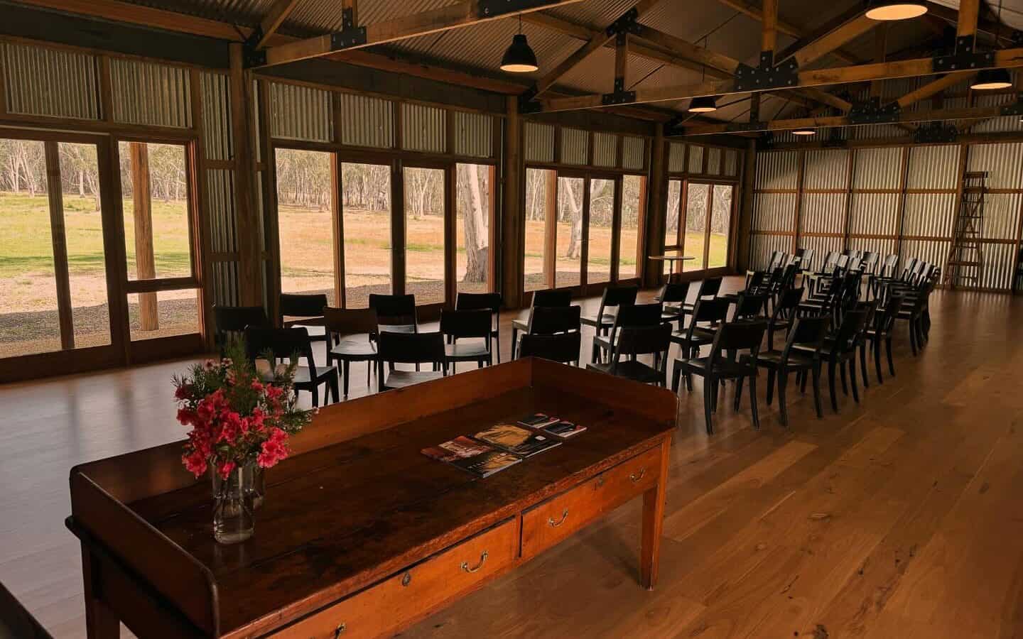 A rustic indoor venue with rows of black chairs facing large windows overlooking a grassy field. A wooden table with books and a vase of red flowers is in the foreground. The space has exposed beams and pendant lights. | Megalong Restaurant