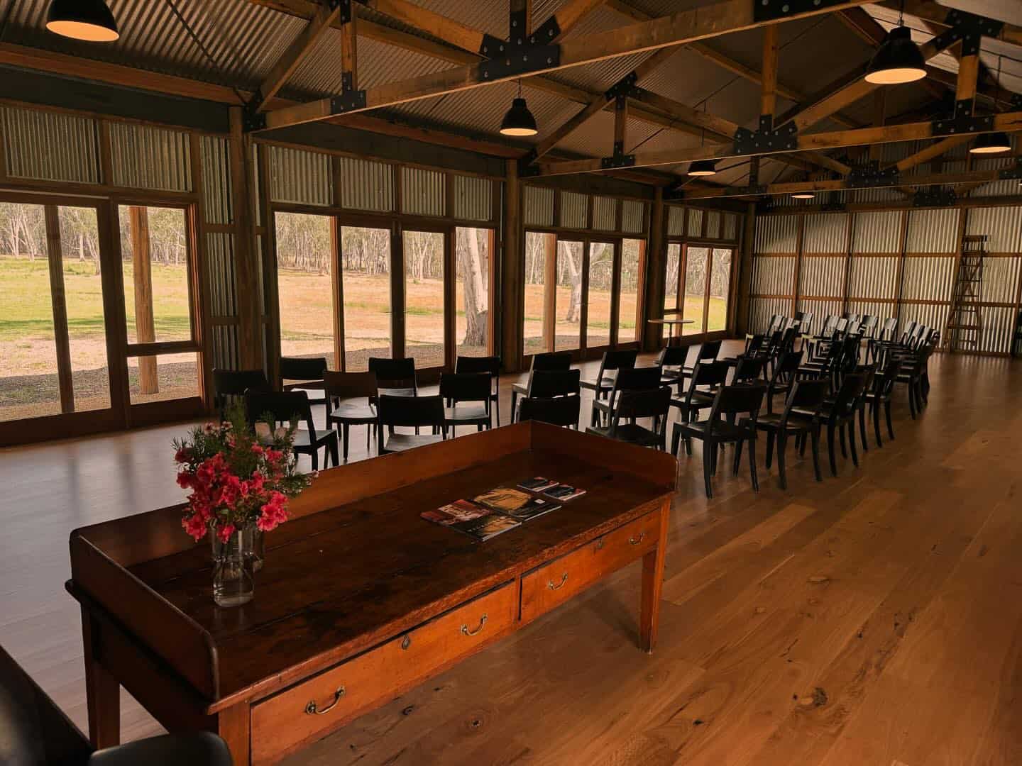 A rustic indoor venue with rows of black chairs facing large windows overlooking a grassy field. A wooden table with books and a vase of red flowers is in the foreground. The space has exposed beams and pendant lights. | Megalong Restaurant