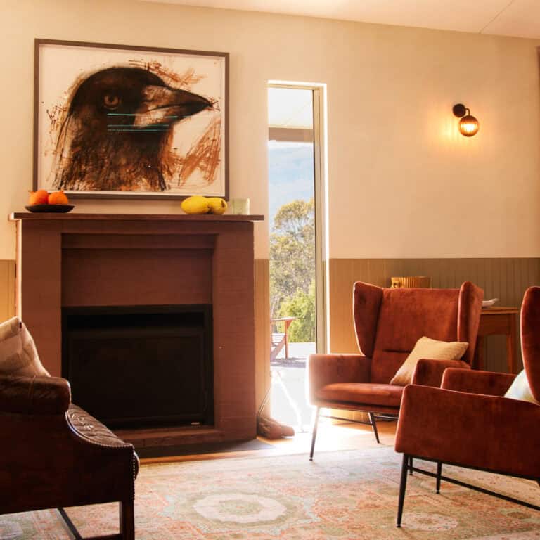 A cozy living room with warm lighting features brown armchairs, a dark fireplace with a bird artwork above, and a window revealing outdoor greenery. A fruit bowl sits atop the fireplace mantel. | Megalong Restaurant