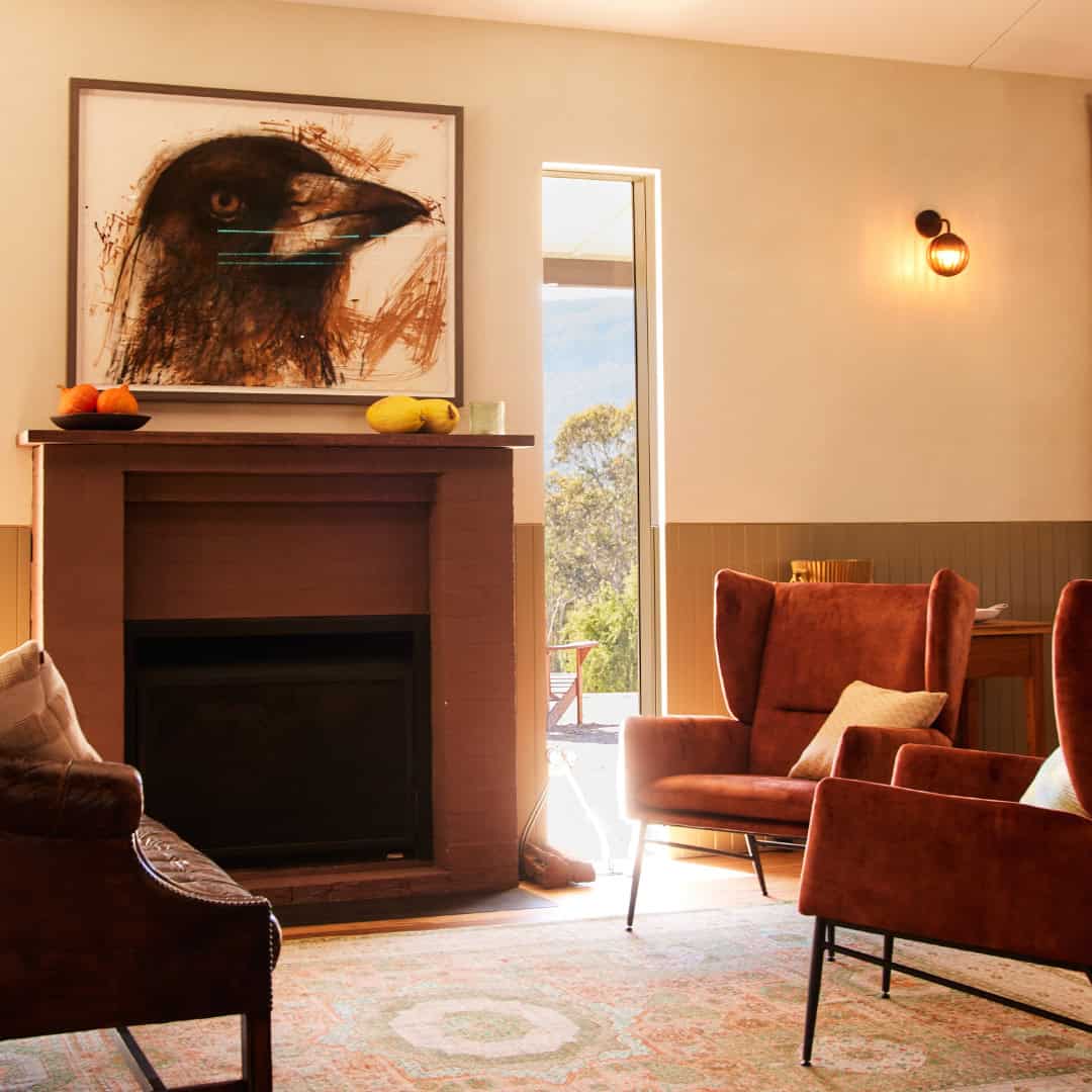 A cozy living room with warm lighting features brown armchairs, a dark fireplace with a bird artwork above, and a window revealing outdoor greenery. A fruit bowl sits atop the fireplace mantel. | Megalong Restaurant