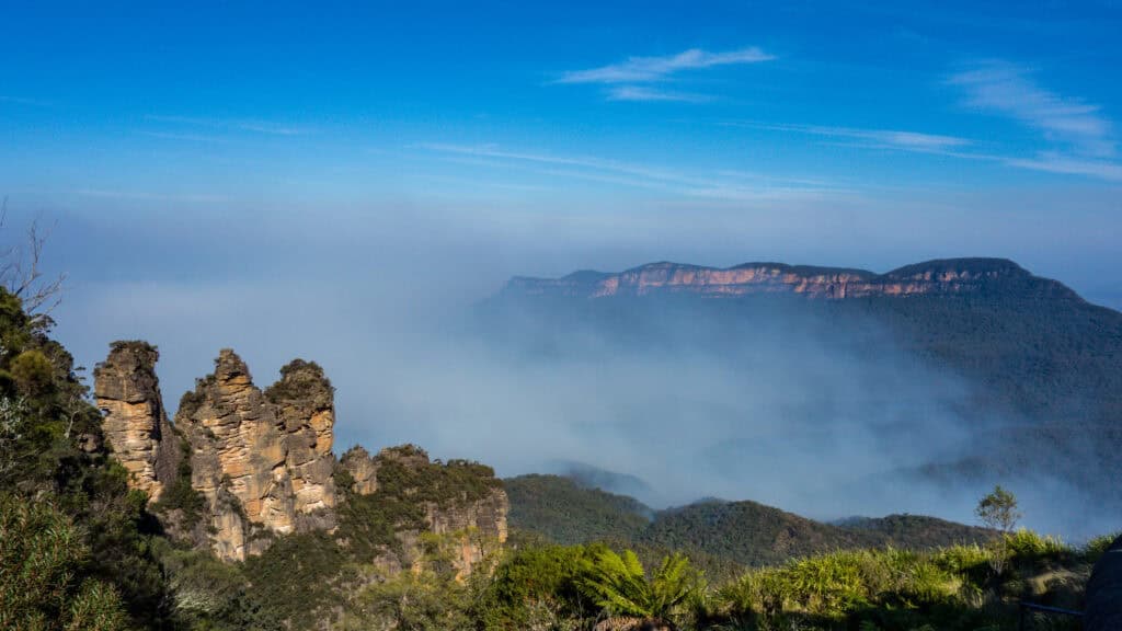 Rock formations known as the Three Sisters rise above lush green vegetation with mist and fog covering the valley, offering one of the best experiences in the Blue Mountains, with distant blue peaks under a clear sky in the background. | Werriberri