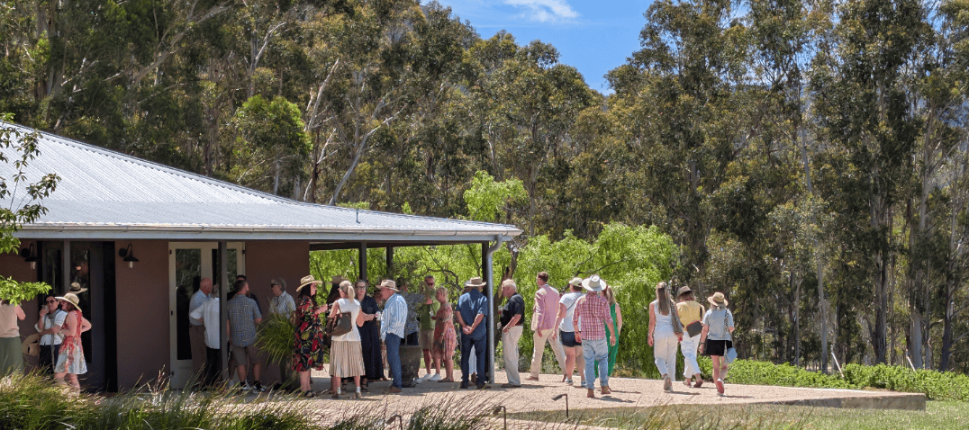 A group of people, many wearing hats, gather outside a house with a metal roof in Megalong, surrounded by trees and greenery on a sunny day for a long lunch celebrating the End of Year 2025. | Megalong Restaurant