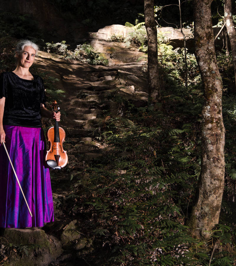 Elderly woman holding a violin in a lush forest environment.