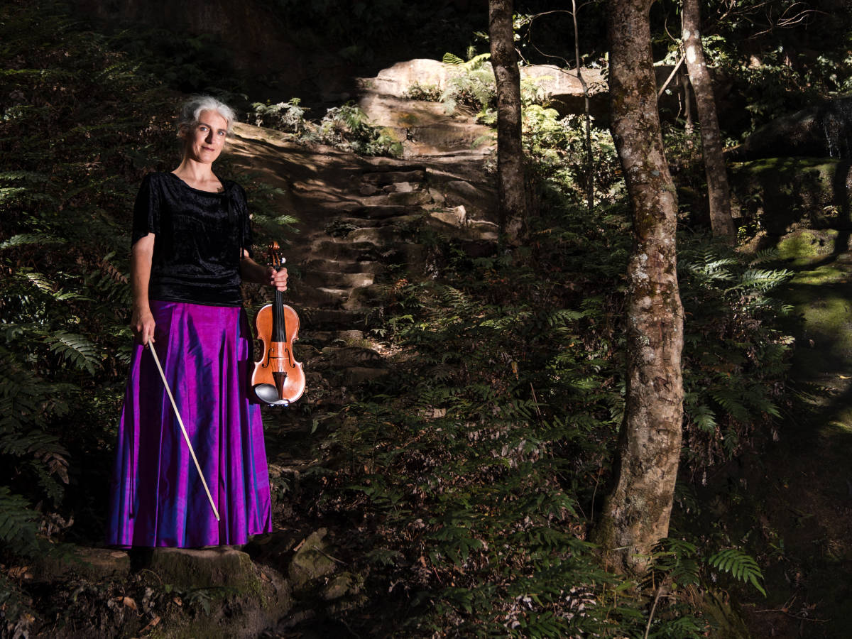 Elderly woman holding a violin in a lush forest environment.