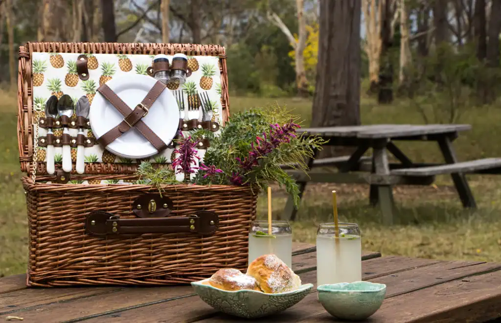 Wicker picnic basket with utensils, drinks, and snacks outdoors.