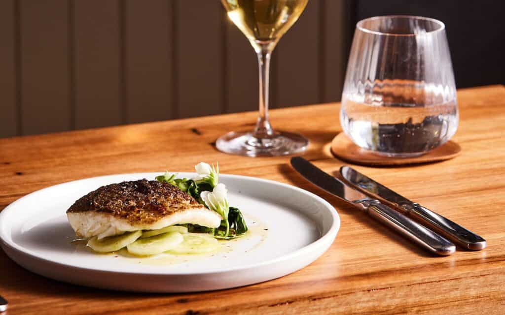 Elegant fish dish with vegetables, wine, and water on a wooden table.