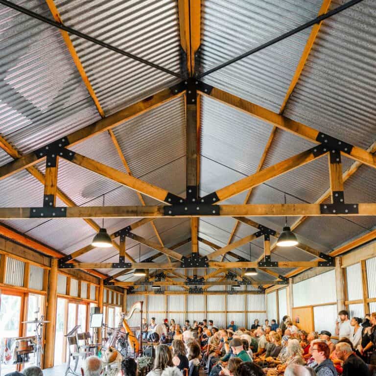 Spacious indoor event space with wooden beams and metal roof, filled with seated audience.
