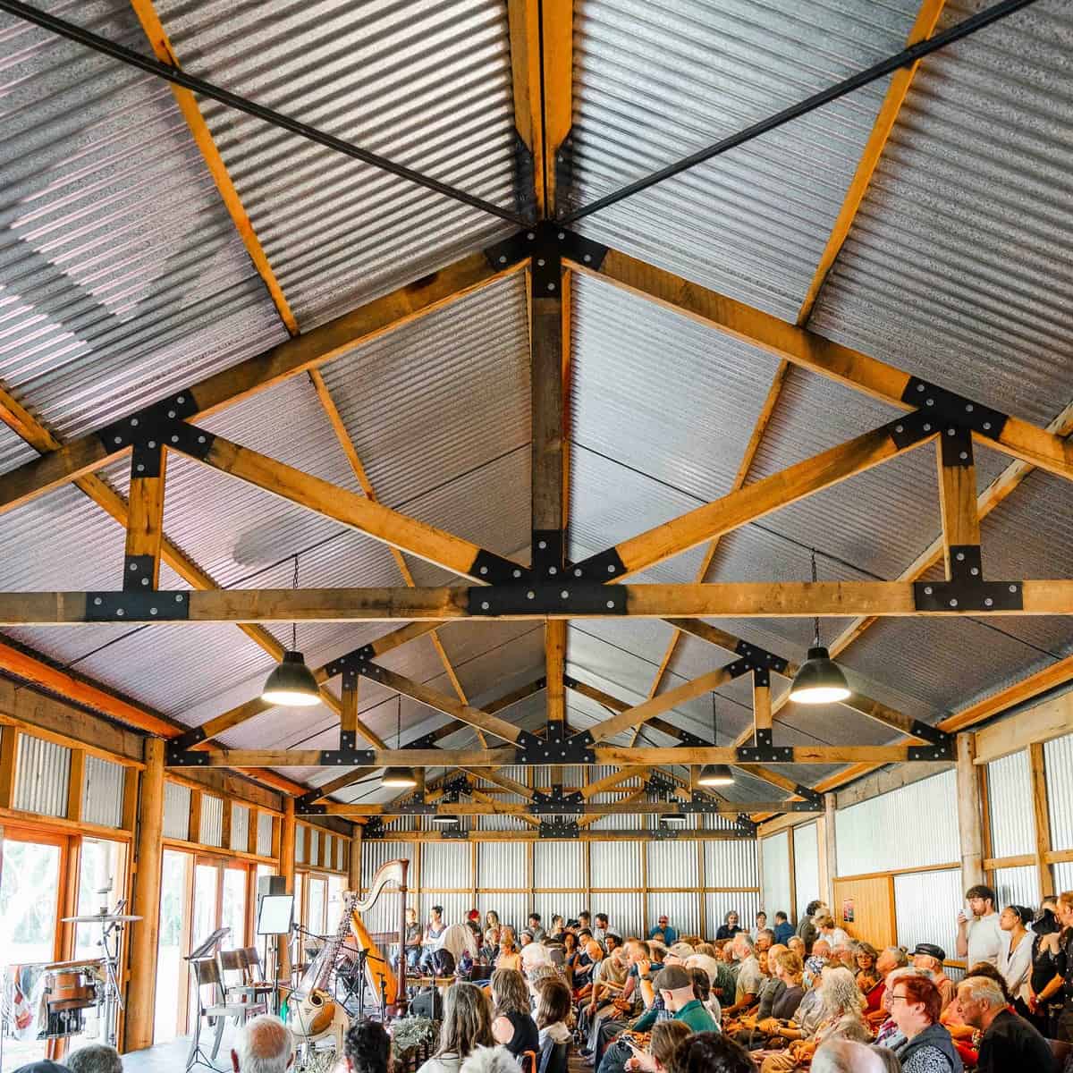 Spacious indoor event space with wooden beams and metal roof, filled with seated audience.