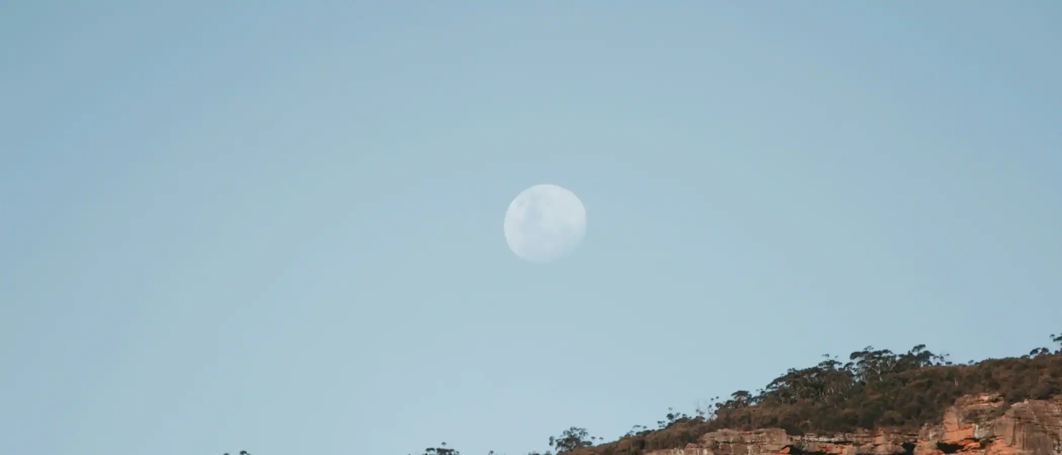 Moon rising over rugged cliffs and forested landscape at dusk.